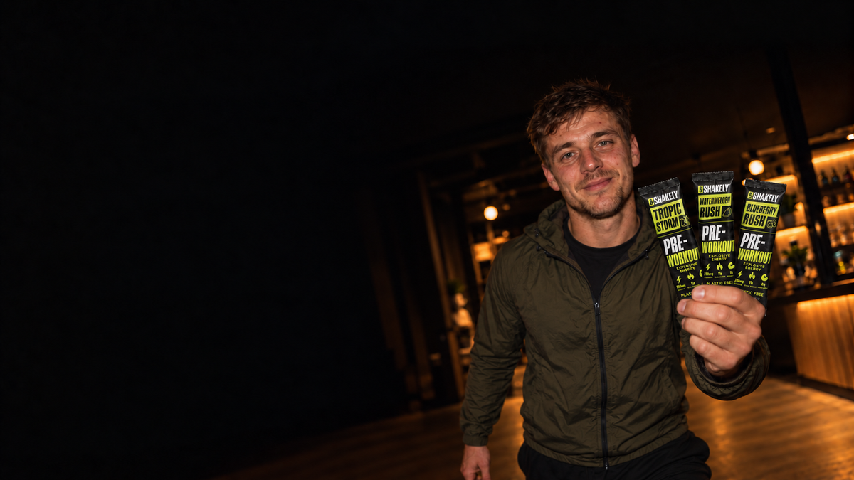Atleet in een donkere olijfgroene jas met drie Shakely-stickpacks in zijn linkerhand, gefotografeerd met flits in een warme avondsetting.